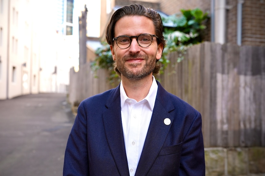 Jason Knauf, with dark floppy hair, wears a blue blazer and white shirt in an alleyway.