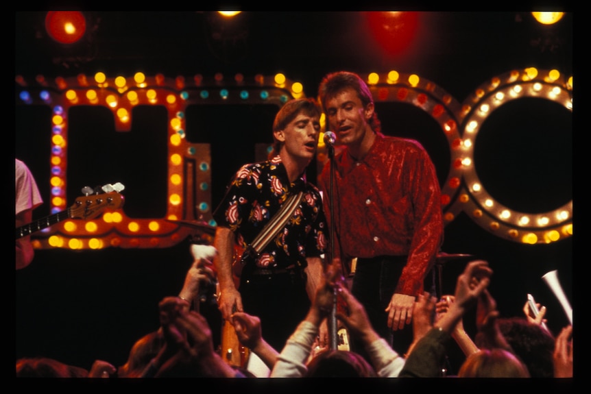Two Australian Crawl band members share a microphone on the Countdown set, illuminated by the show's lit-up logo.