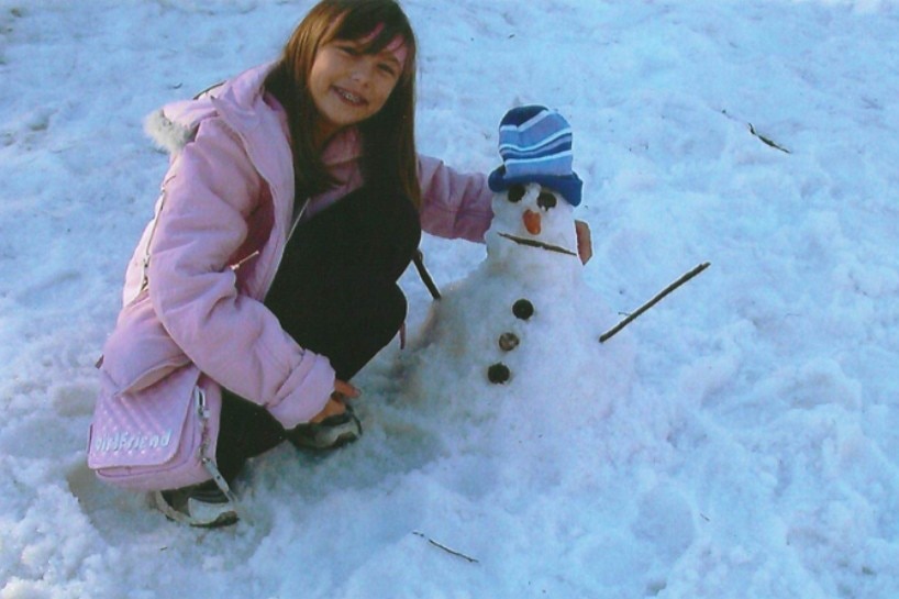 A young child in a pink puffer jacket crouching in the snow next to a snowman.