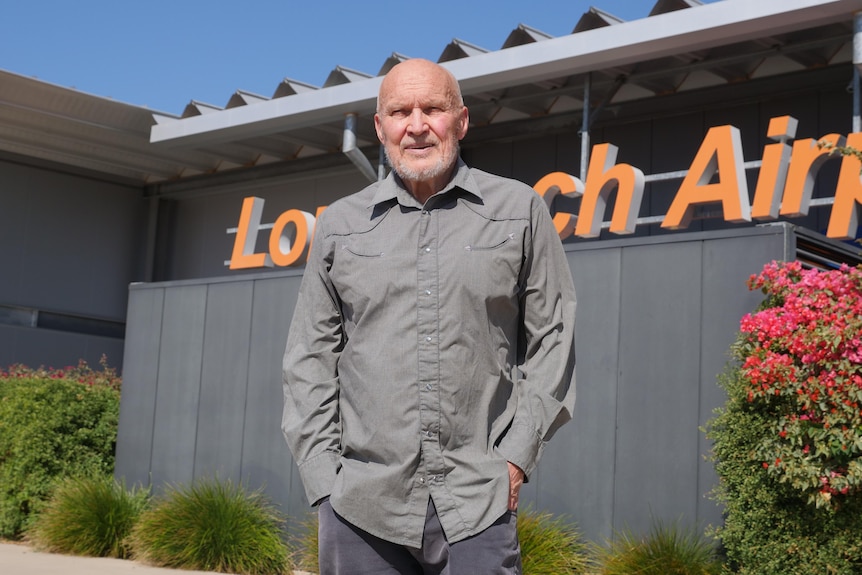A man wearing a long-sleeved outback workshirt has a stern expression, standing in front of a "Longreach Airport" sign.