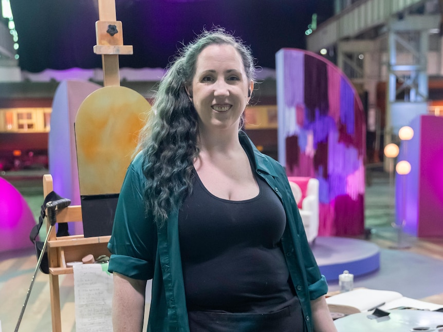 A woman with long brown curly hair smiles at the camera. Behind her is an easel
