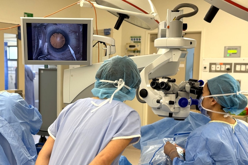 Medical staff stand around a patient, with the patient's eyeball displayed on a screen to guide the surgery.