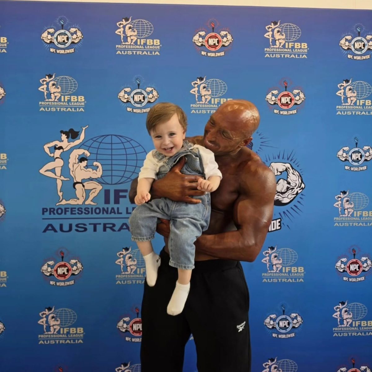 bodybuilder holding son in front of IFB banner
