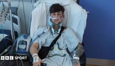 Liam McCracken, who is wearing a patterned hospital gown, is sitting in a chair next to a hospital bed. He is wearing a neck brace and is surrounded by medical equipment.