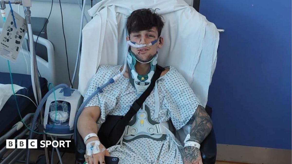Liam McCracken, who is wearing a patterned hospital gown, is sitting in a chair next to a hospital bed. He is wearing a neck brace and is surrounded by medical equipment.
