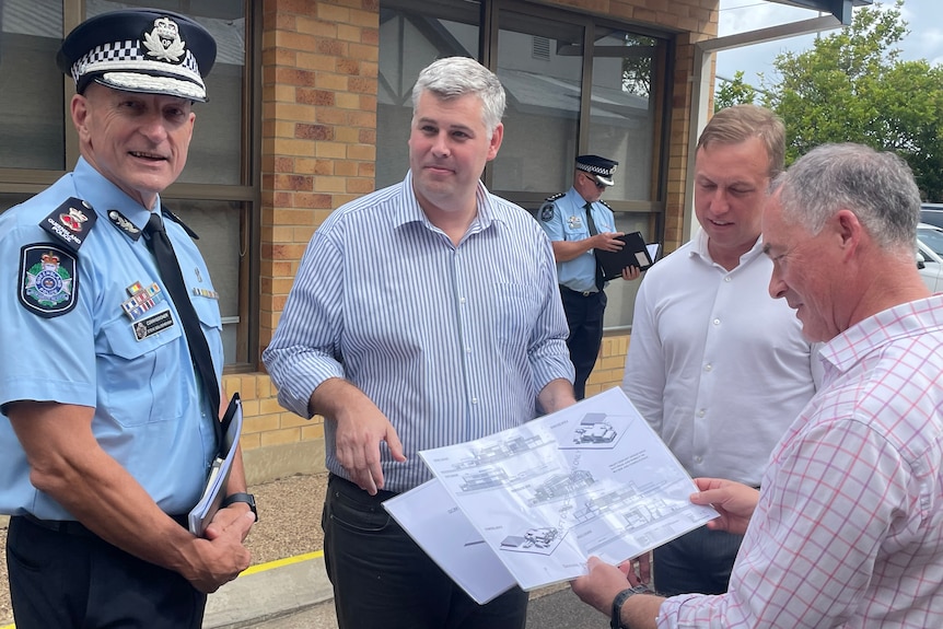 Photo of four men, one is a police officer, three are politicians. One politician is looking at a map