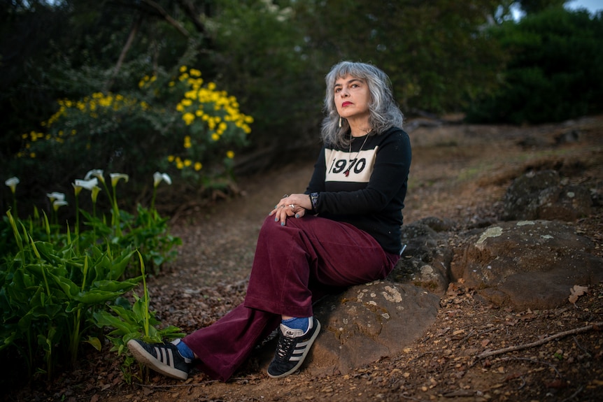 A woman sits on some rocks in a black jumper with "1970" printed and yellow and white flowers in the background.