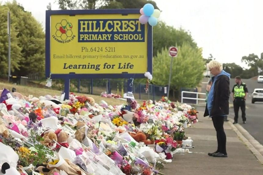 Man stands on footpath before a pile of floral tributes outside a primary school, sign reads 'Hillcrest Primary School'.