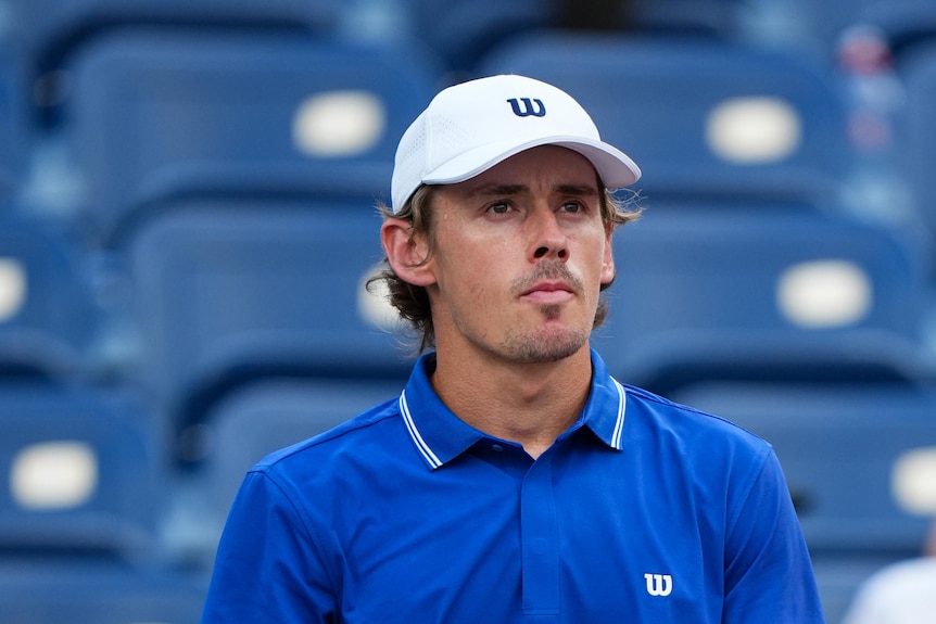A male tennis player in a blue shirt sits courtside, looking frustrated.