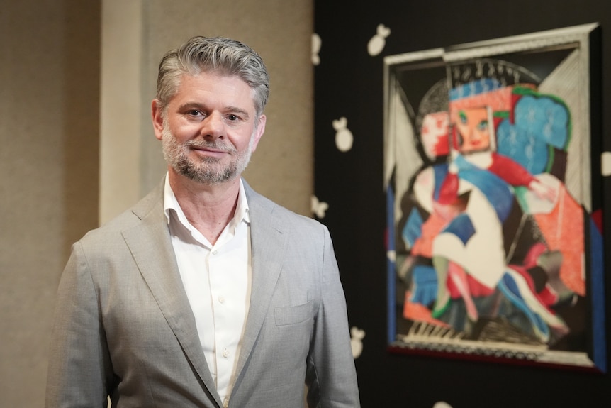 A man with short grey hair wearing a grey suit stands in an art gallery in front of a cubist portrait.
