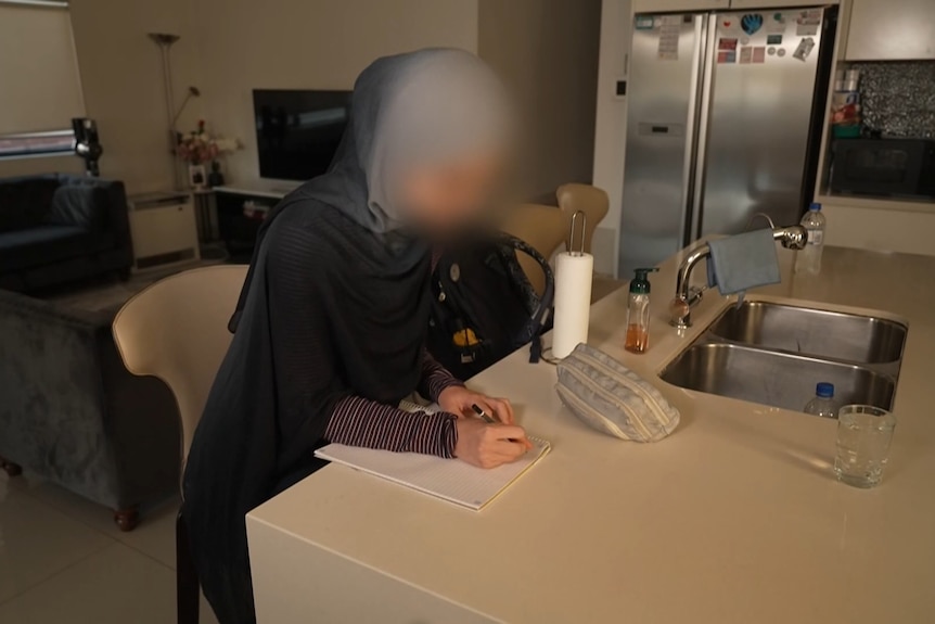 Janna, wearing a hijab, sitting at a kitchen bench writing in a note pad.
