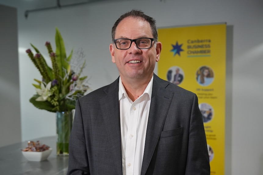 Greg, with short dark hair and wearing a grey suit, stands in an office smiling.