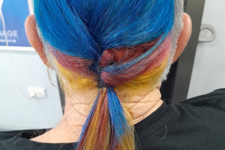 The back of a man's head with long hair dyed red, blue and yellow in a pony tail.