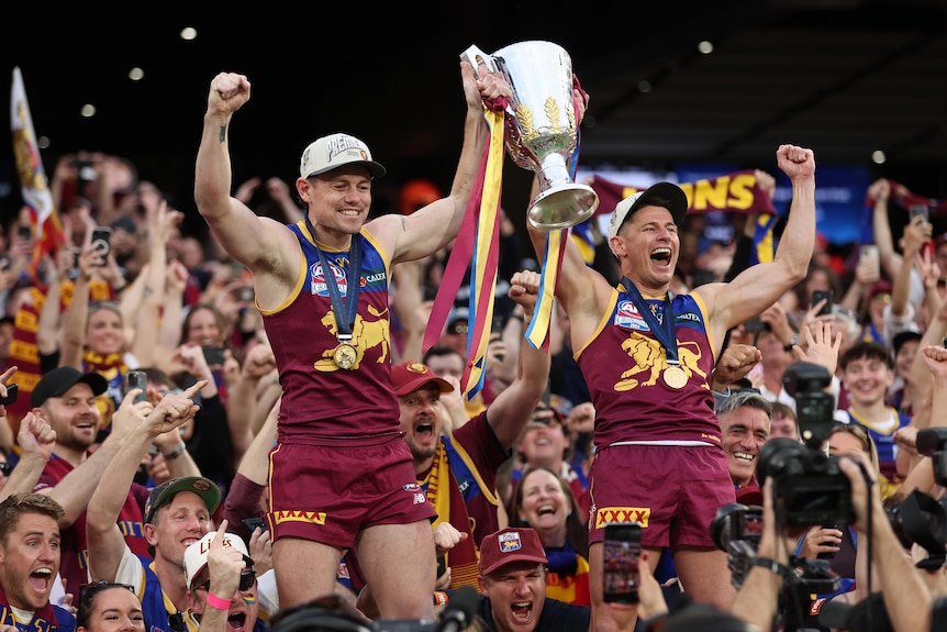 A pair of AFL players hoist a trophy in triumph in front of a screaming stadium crowd.