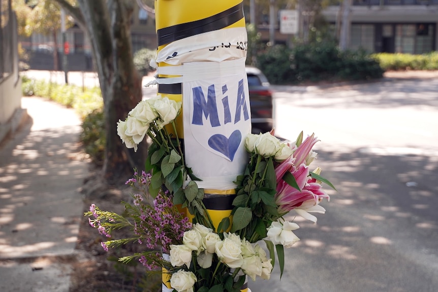 flowers with a sign that read mia on a pole at the site where a young police  officer died after the car crashed into a tree