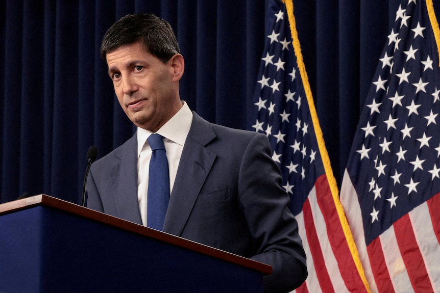 A man in a suit and tie speaking at a podium in front of two US flags.