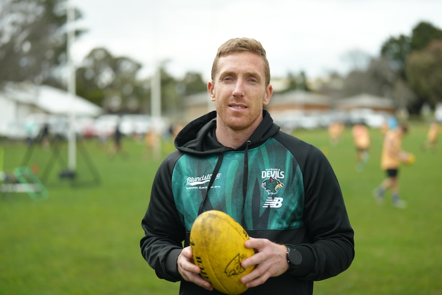 A man wearing a black and green hoodie smiling at the camera holding a yellow football.