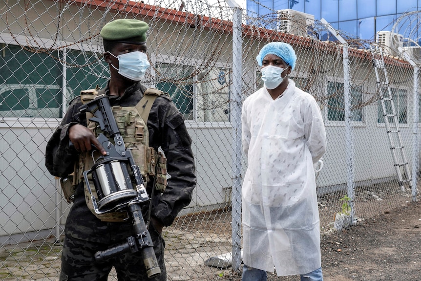 A man in military uniform standing guard in front of another in PPE.