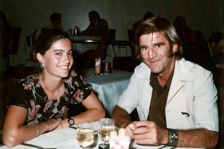 A pretty young woman and a man in a safari suit sit at a restaurant table with wine glasses.