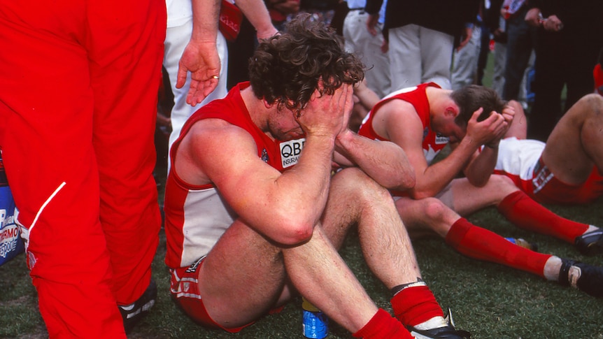 Two Sydney Swans AFL players slump on the ground with their heads in their hands after a grand final loss.