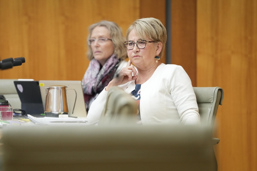 A woman with short blonde hair and glasses leans back in a sage green office chair resting her head on her right hand.