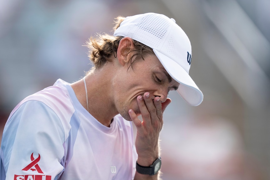 Alex de Minaur rubs his face with his left hand while wearing white cap