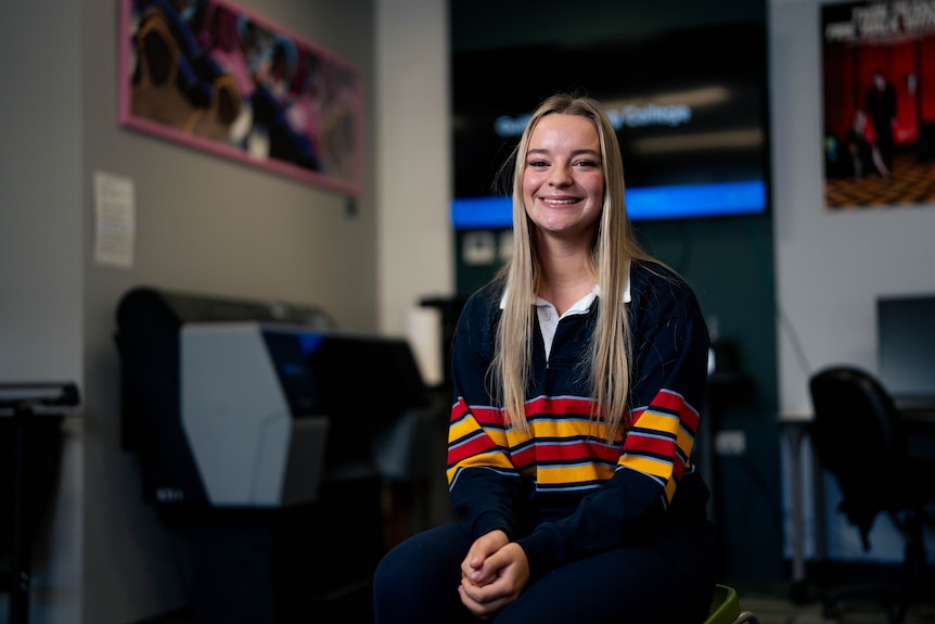 Lacey sits on a chair in a classroom with a printer and TV behind her, wearing a school rugby jumper and smiling.
