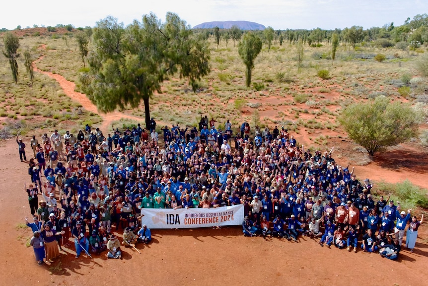 A large crowd of people gathers together in the desert. Some at the front hold a banner saying IDA.