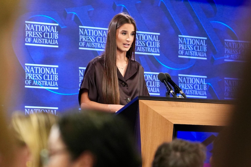 Chanel Contos, a young woman with brown hair, speaks at a microphone on a stage.