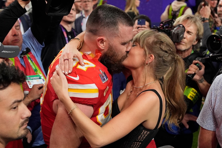 A muscular footballer kisses a blonde pop star as media and camera surround them