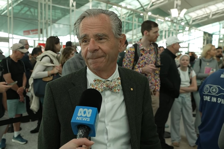 Con smiles in a suit, bow tie, has grey hair, and speaks into a blue ABC microphone.