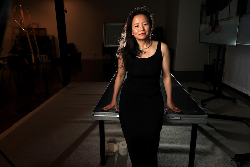 Woman with dark long hair wears a black maxi dress leaning with her hands on a table.