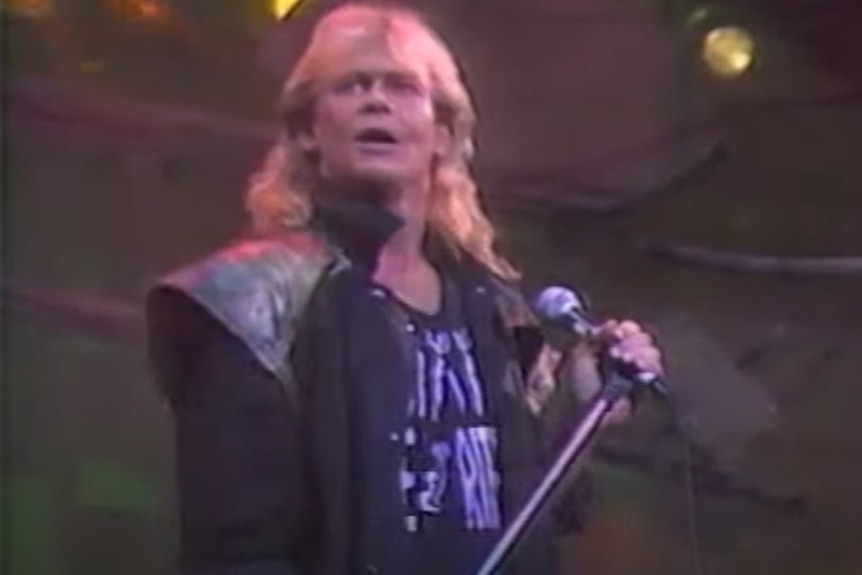 A man with long blonde hair wearing a big black jacket holds a microphone.