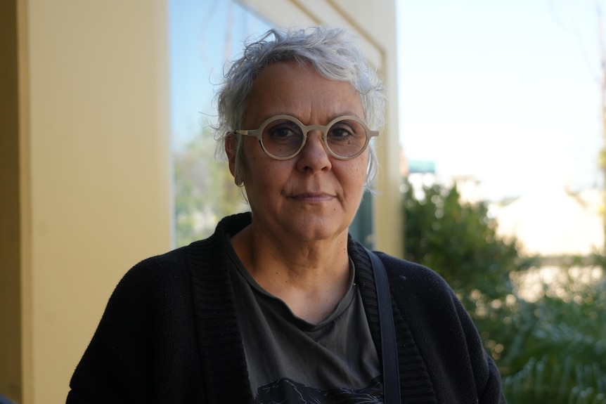 Ms Best with short white hair wearing glasses and a black jumper and shirt looks serious with plants behind her.