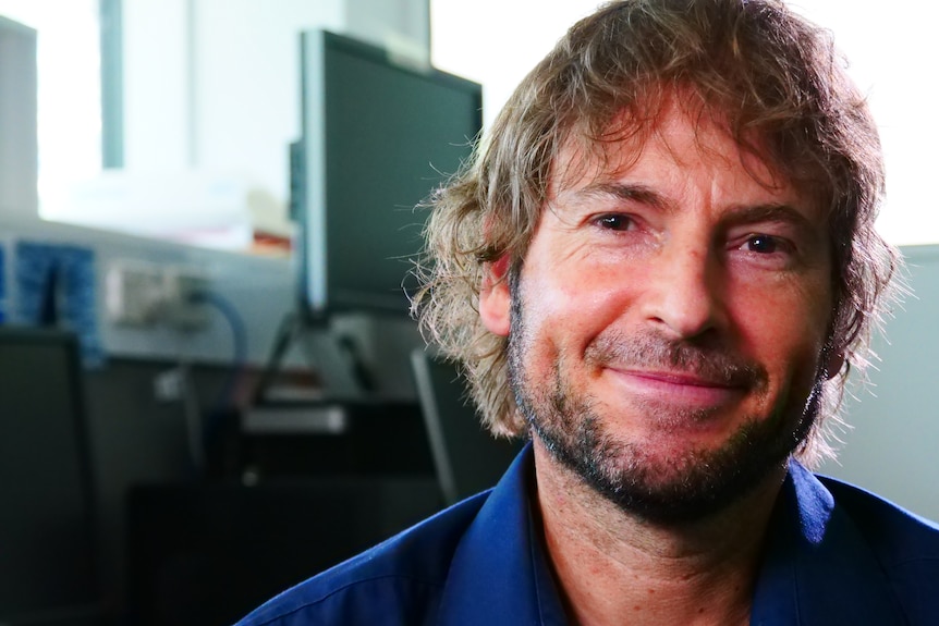 Man smiles. He wears a blue shirt and has wavy, fair hair.