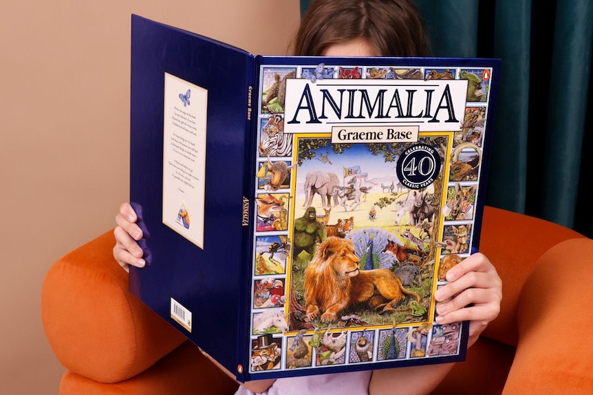 A child sitting on an orange armchair, holding a copy of Animalia by Graeme Base in front of their face.