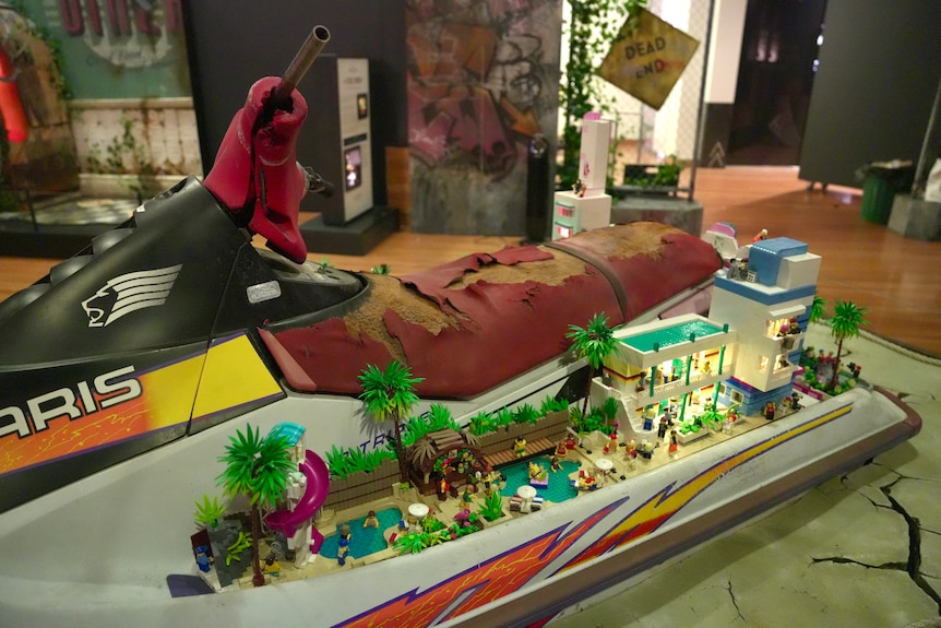 A jet ski with a worn-out seat, a lego hotel is where one would put their foot, with pools, a water slide and palm trees.