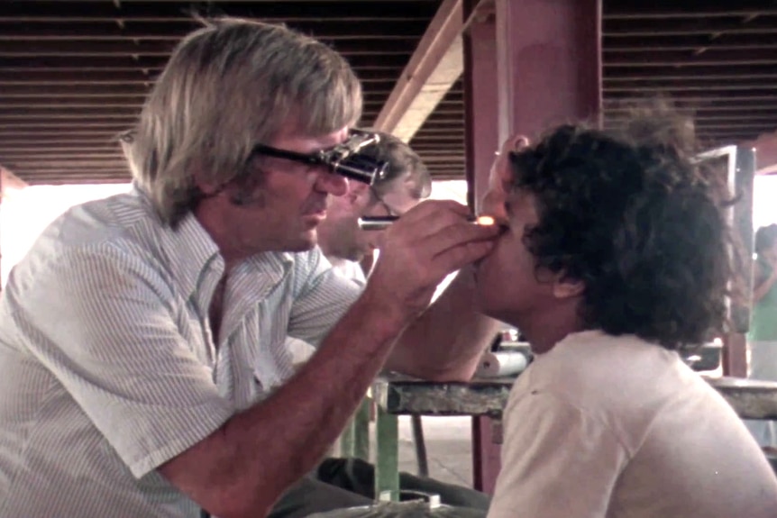 A man wearing ophthalmological glasses instects the eys of a young Aborginal boy.
