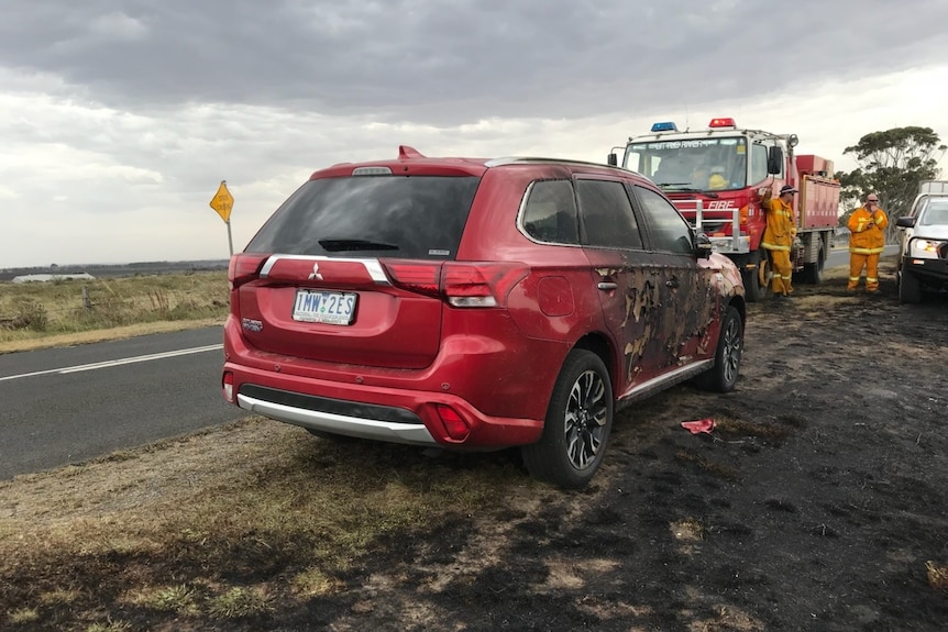 A burnt out EV car next to a fire truck with firefighters nearby