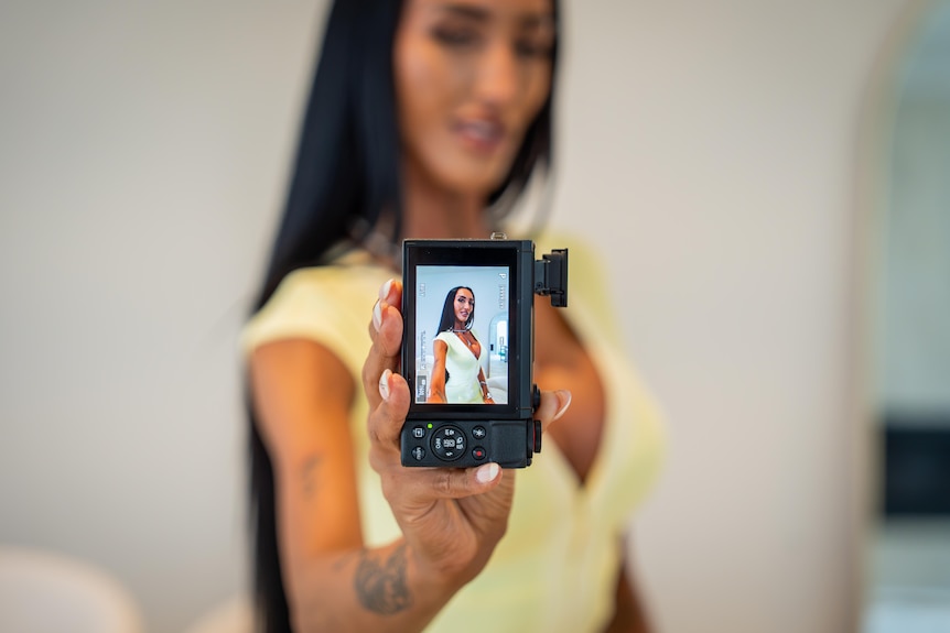 A woman in a yellow dress holds up a camera with a photo of herself displayed