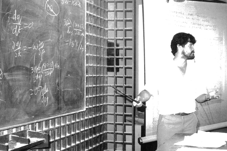 Black and white photo of John McBride standing near a blackboard.