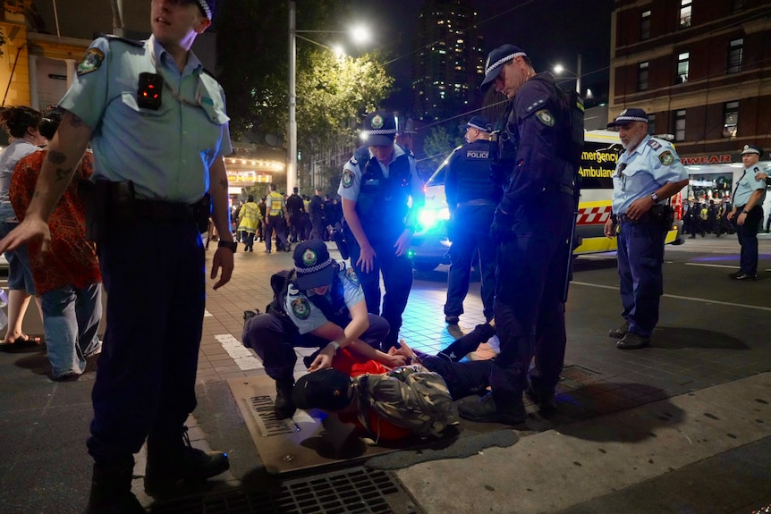 Police stand over m an on the ground in the dark, lights of ambulance behind