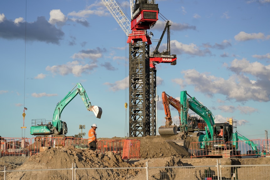 cranes and bulldozers on a building site there is a worker in high vis in the foreground standing next to piles of dirt
