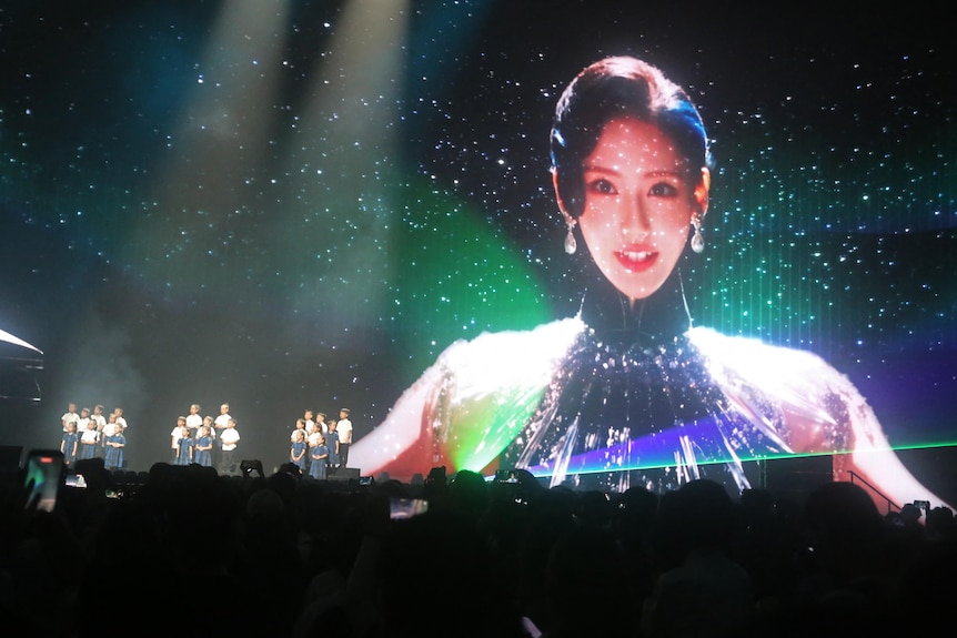 The image of a woman wearing large earrings and a black dress appears on a large screen behind a stage.