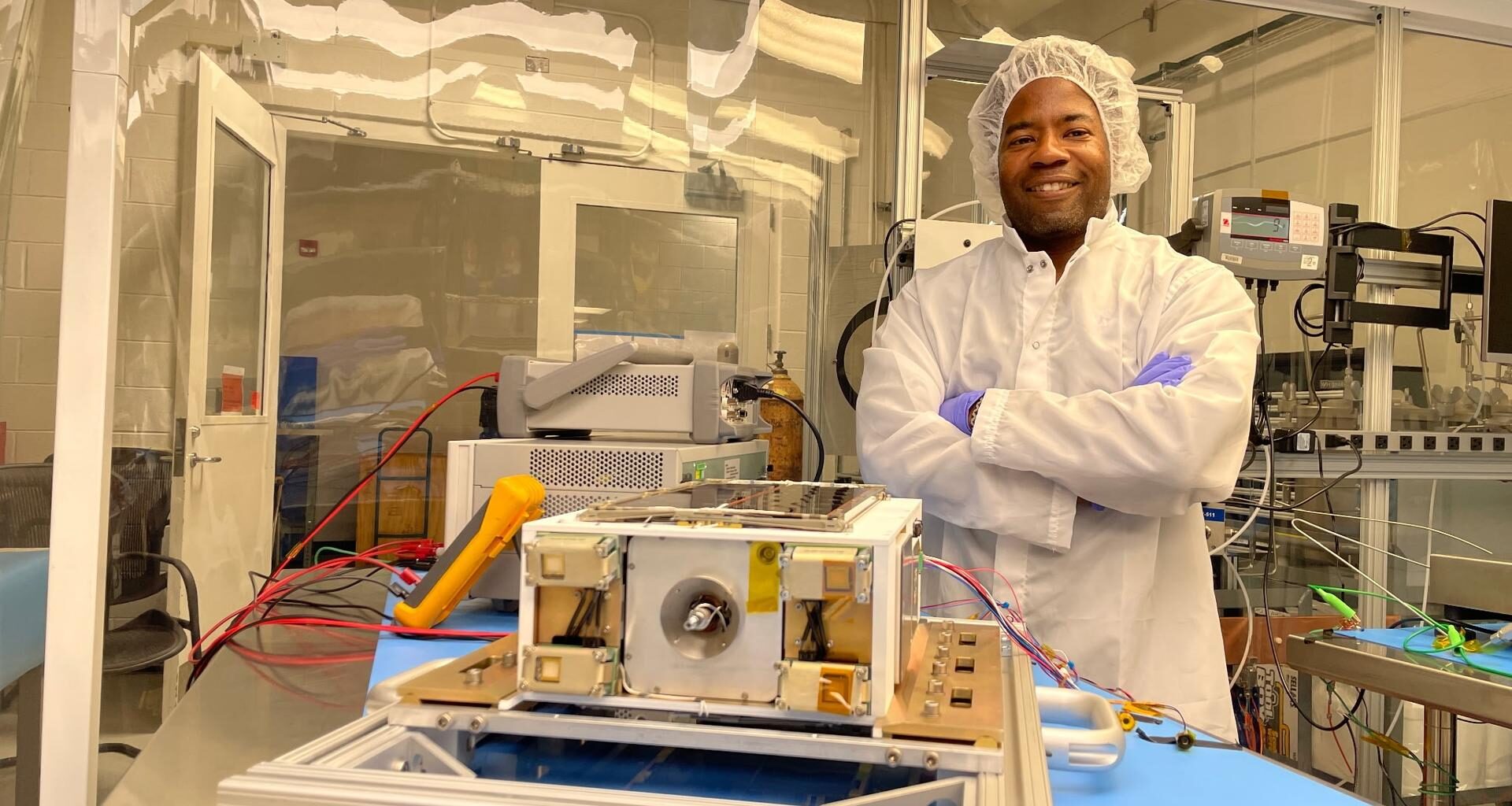 An Album of photos showing NASA engineers and technicians in white protective clean room suits working on a small, rectangular spacecraft module. They are shown inspecting the hardware on a laboratory workbench, wrapping it in silver foil, and loading it into a large, metallic vacuum chamber at NASA's Marshall Space Flight Center