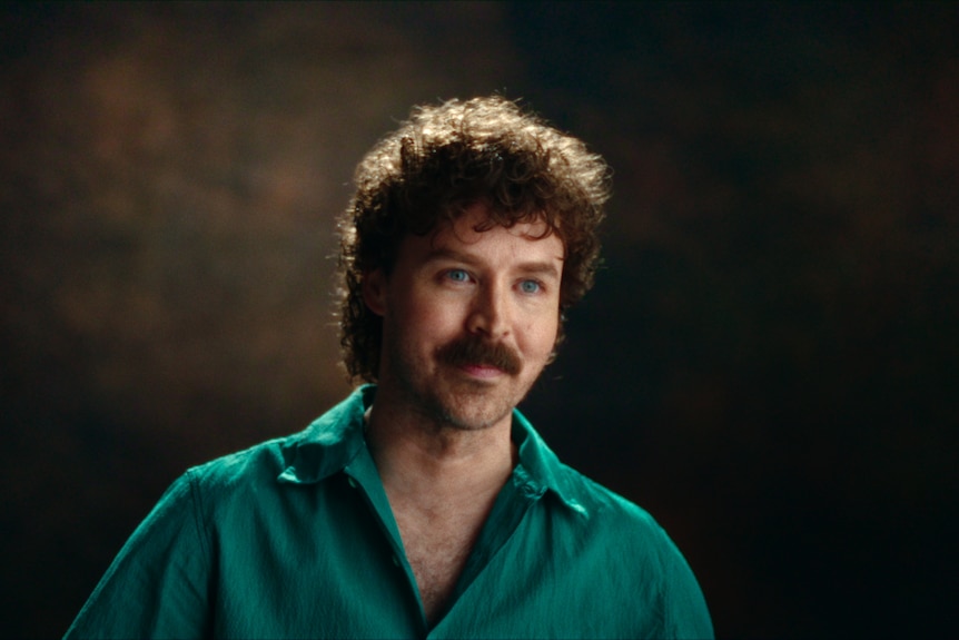 A white man with curly brown hair and a big moustache wearing a green shirt