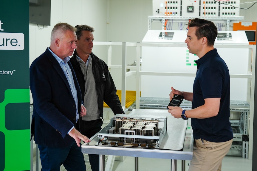 Jeremy Rockliff stands in front of computer processing units, speaking with two men.