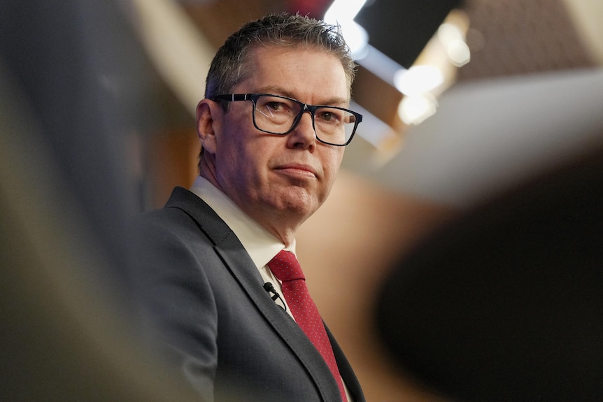 Pat Conroy, a white middle aged man with red tie and glasses, staring as camera captures glimpse through blurred foreground.