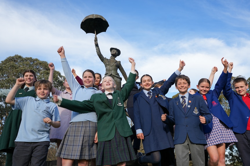 A group of schoolchildren in various different school uniforms cheer in front of a bronze statue of Mary Poppins.