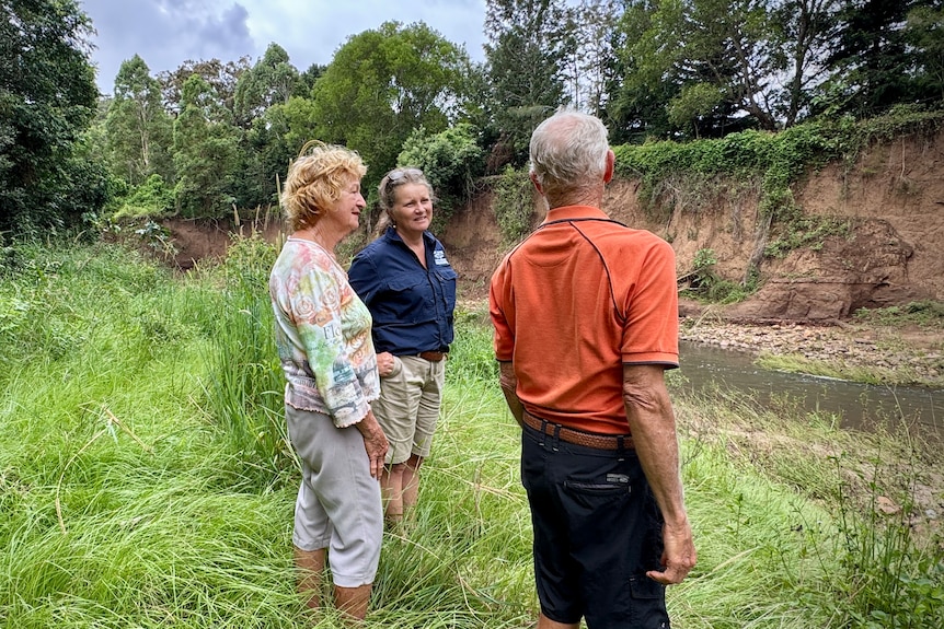 Three people stand across a creek with a badly eroded bank.
